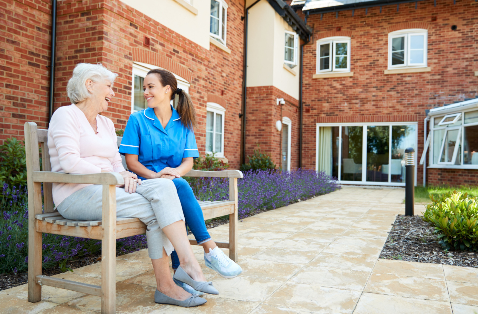 a caregiver and a senior woman sit outside a senior living community while chatting and smiling