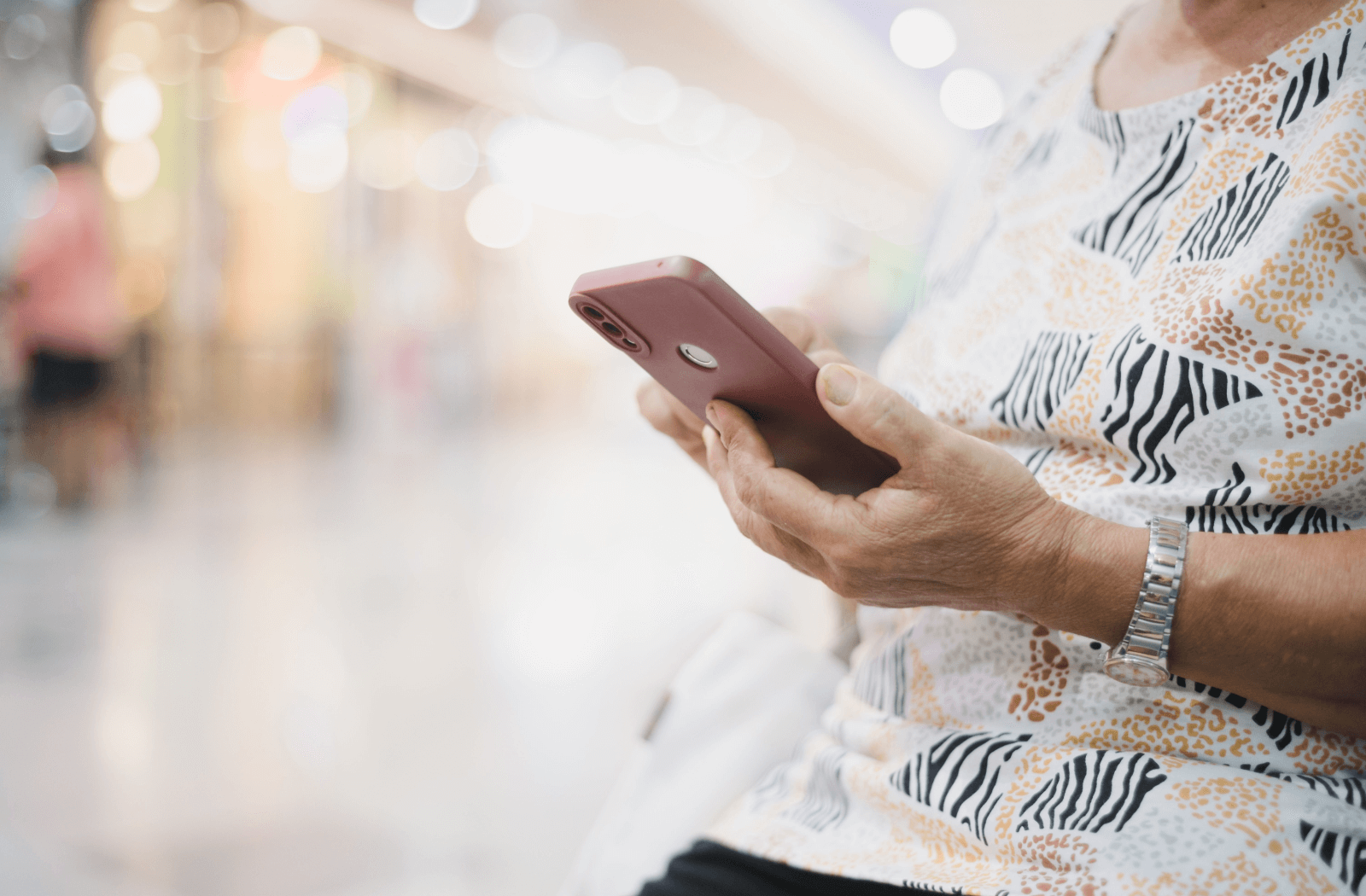 A close-up of a senior person holding an iPhone in a maroon case against a brightly lit background.